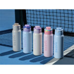 row of five pastel-colored water bottles with flip lids lined up on a blue tennis court beside a net, casting long shadows.