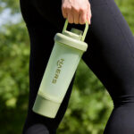 light green sports shaker bottle being carried by a hand outdoors with greenery in the background, worn black leggings visible nearby.