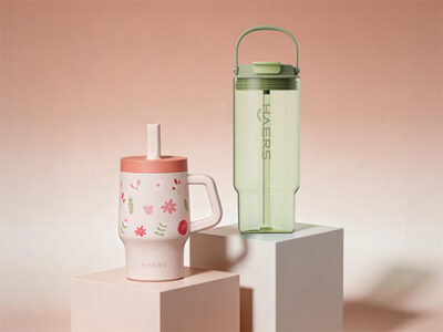 pink floral insulated mug with straw and a light green water bottle with a handle on display pedestals against a soft pink gradient background.