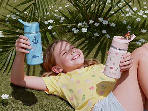 smiling girl lying on grass in a yellow shirt, holding a blue bottle with an anchor and a pink bottle with a butterfly on the cap.