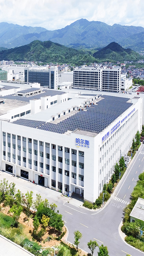 aerial view of a white multi-story office building with solar panels on the roof, haers signage, surrounded by roads and green hills in the distance.